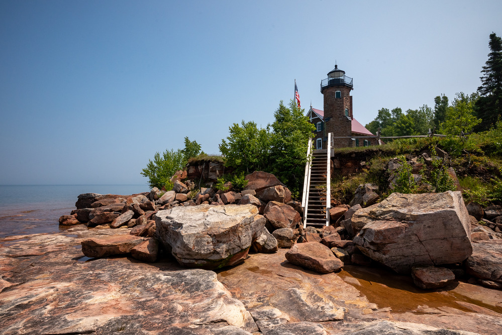 Brick lighthouse perched above a rocky Lake Superior shoreline, with a staircase leading down to the shore.