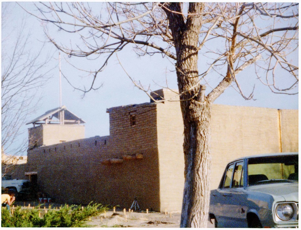 A car is parked next to a tree a short distance to the north of the reconstructed fort structure. 