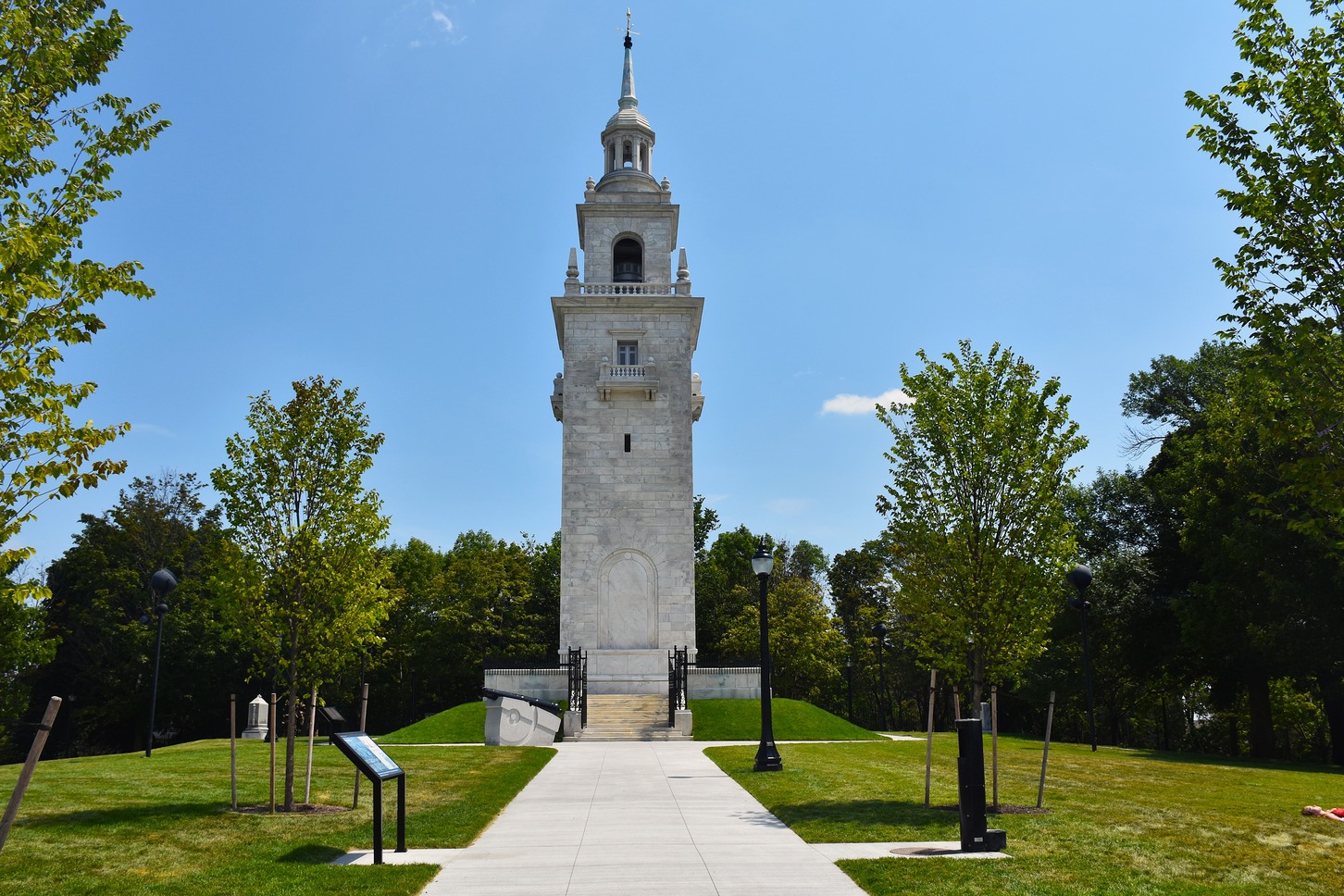 A paved path in grassy grounds leads to a tall granite monument with spire