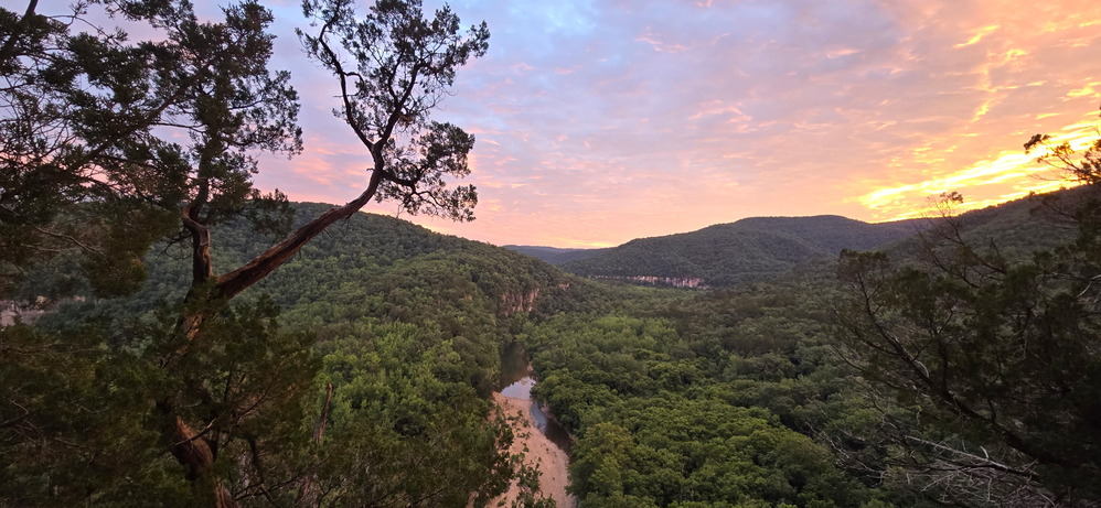 A pink and orange sky over the river surrounded by green trees.