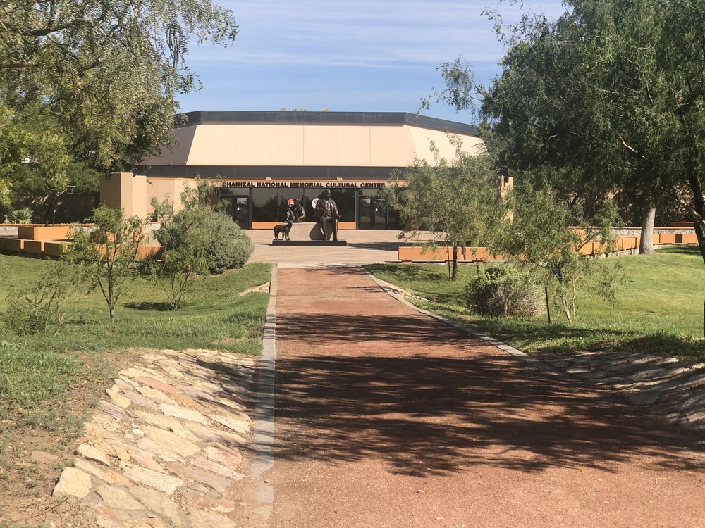 A packed red gravel path in foreground is lined with trees and a grassy area. It leads to an esplanade and sand colored two story building in the background. In the middle of the esplanade is a sculpture with multiple figures.