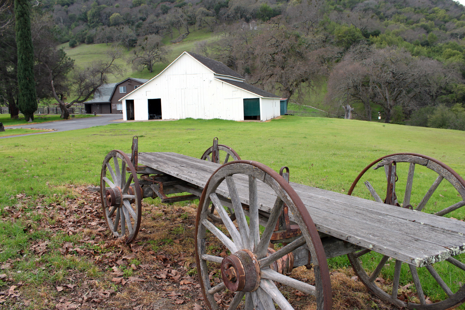 An old bard and a wagon wheel cart sit amongst trees, grass and hills. 
