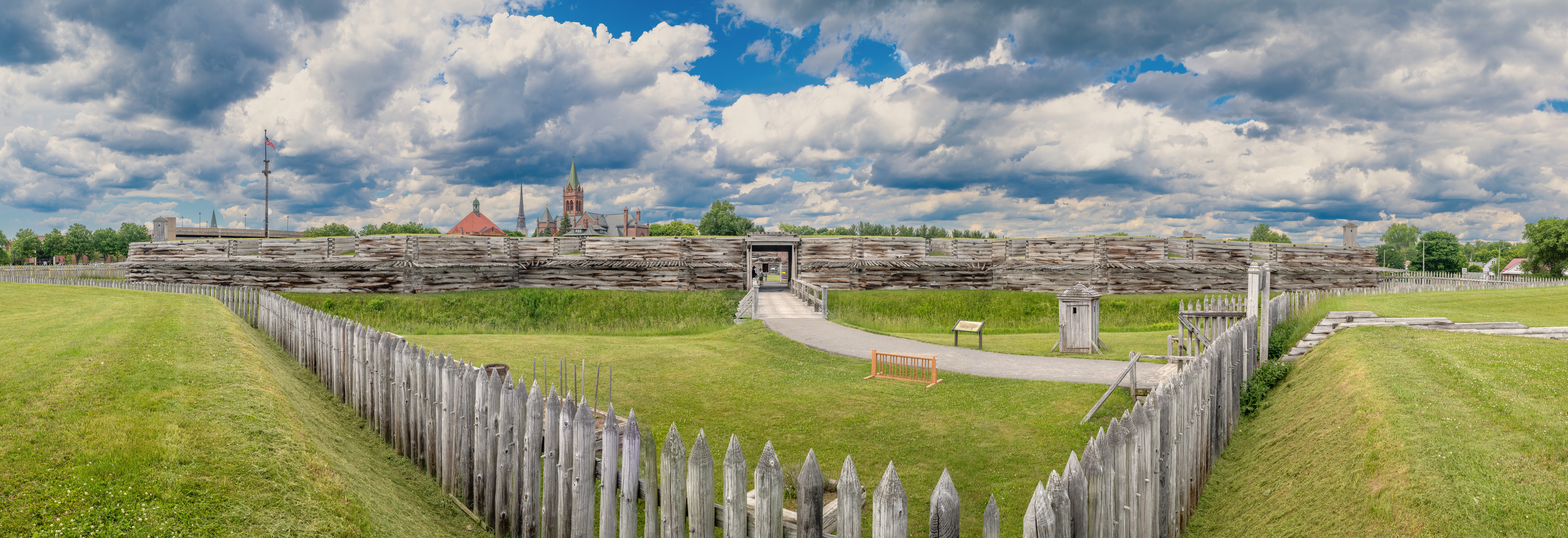 A large wooden wall stretches in front of you. A trail and wooden bridge lead inside of it. 