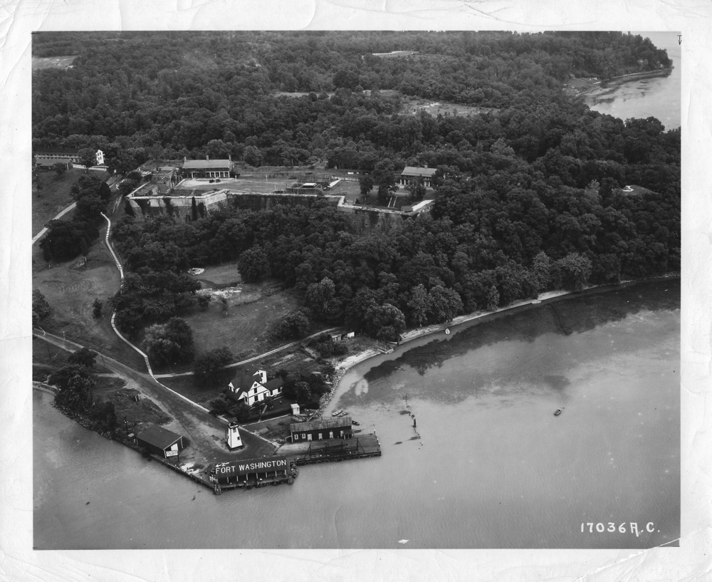 Photo shows the dock and lighthouse area.  The lighthouse keepers quarters is behind the lighthouse.  The fort is surrounded by heavy tree coverage, with several trees growing out of the southwest demi-bastion.  A small area of PIscataway Creek can be seen to the right of the photo.