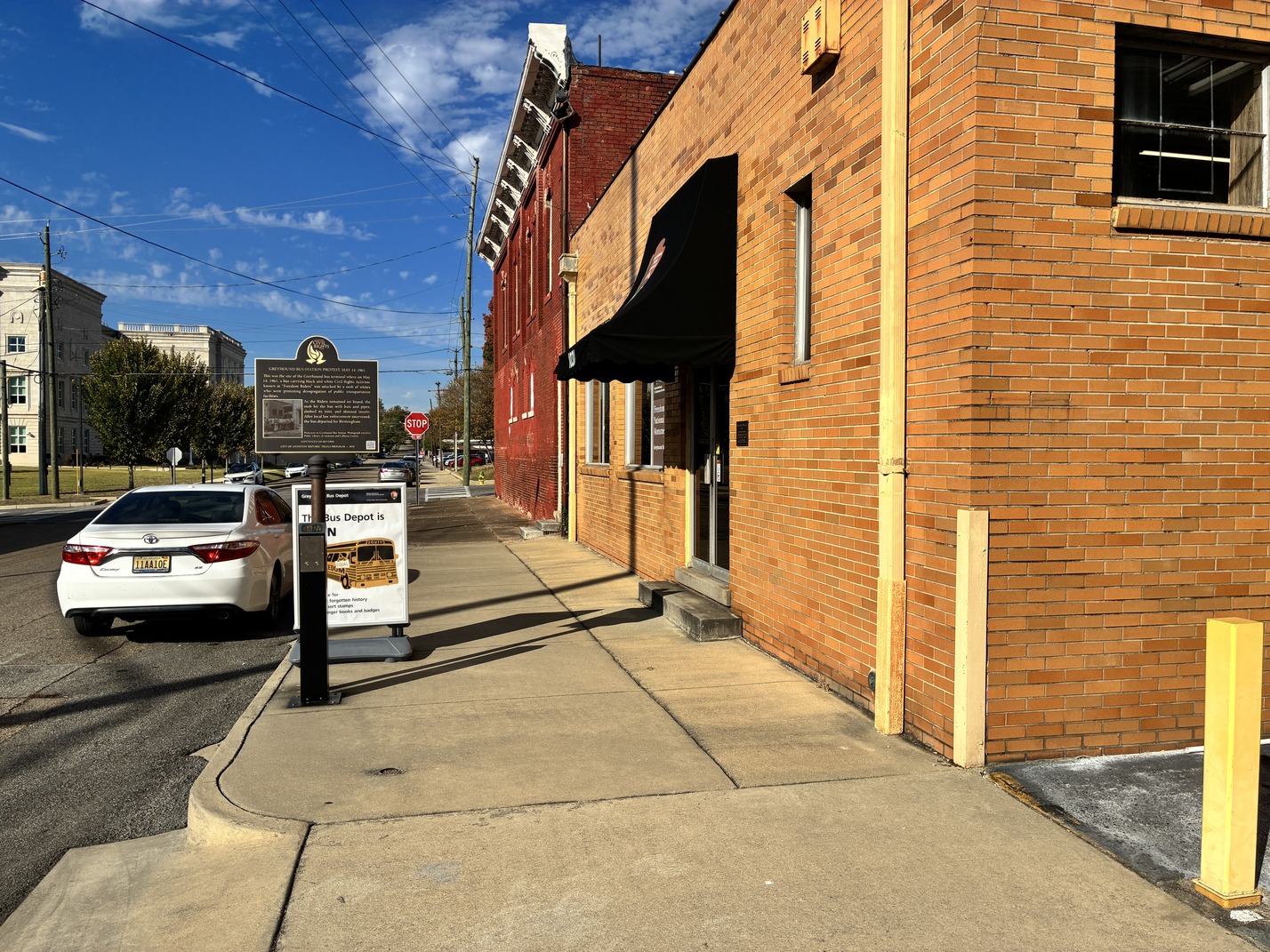 A tall, one-story brick building separated from the street by a sidewalk. A metal historic marker is posted at the edge of the street. 