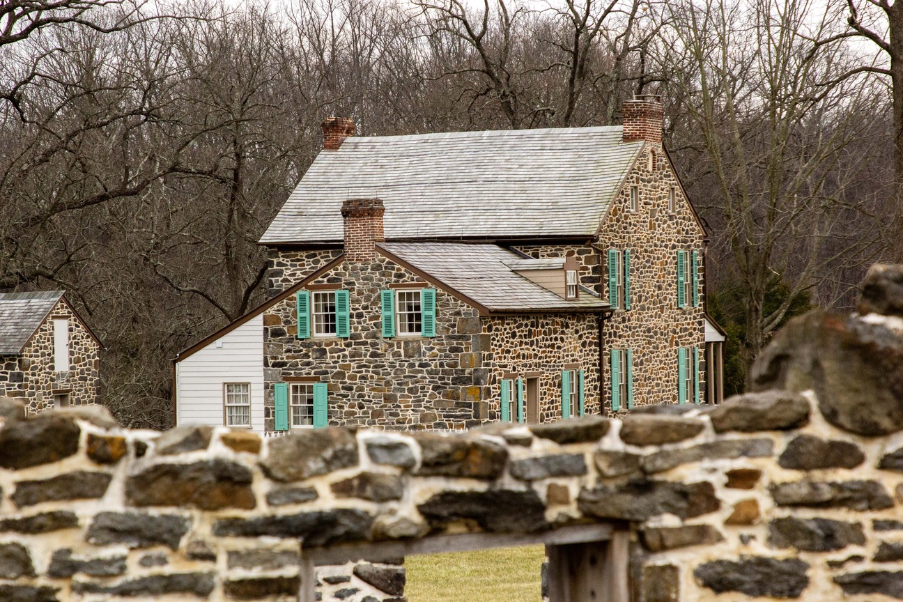 A two-story stone farmhouse with green shutters. Behind the farmhouse is a wall of bare trees. In the foreground are the remains of a stone wall. 