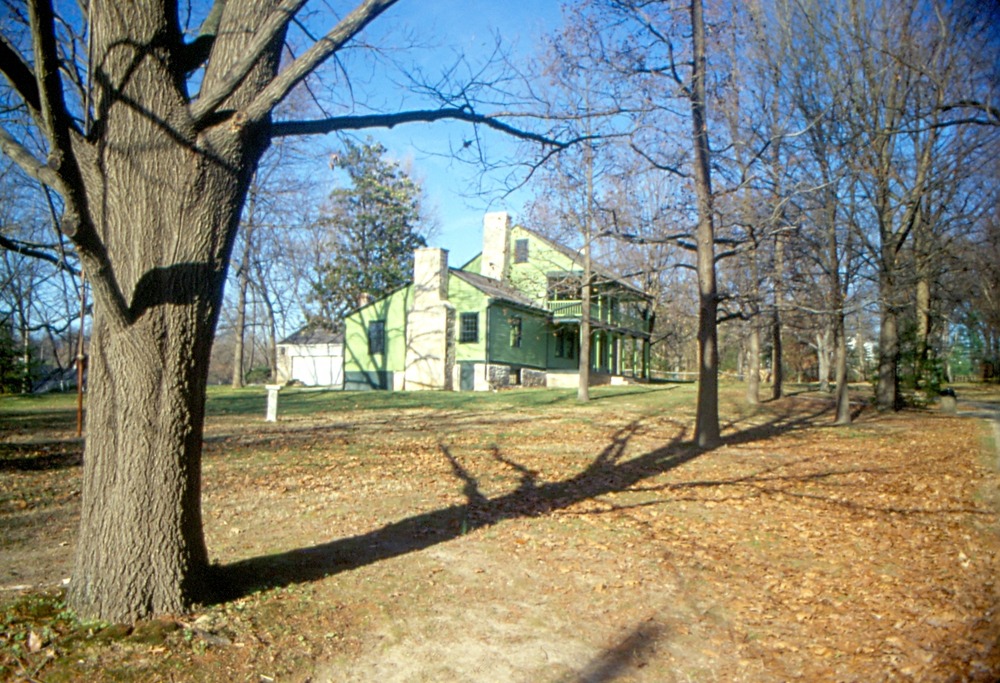 Image of a distant view of a green house with a two-story porch across the front and a single-story wing.
