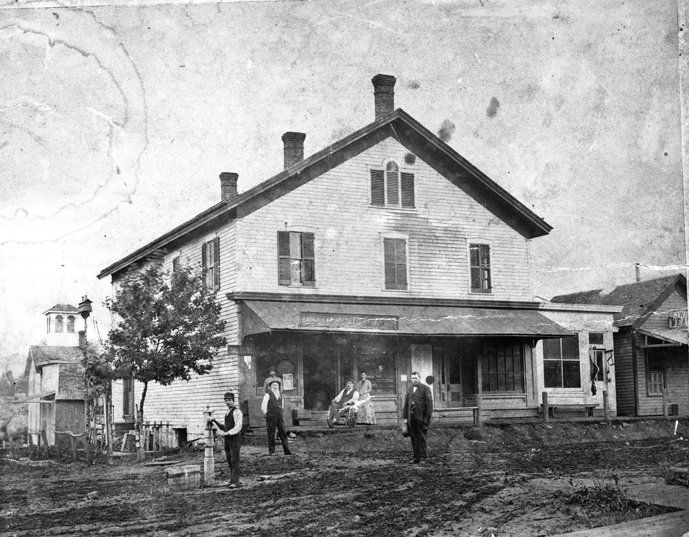 A gabled three story building occupies a downtown corner where people in 1800s dress stand by a water pump.