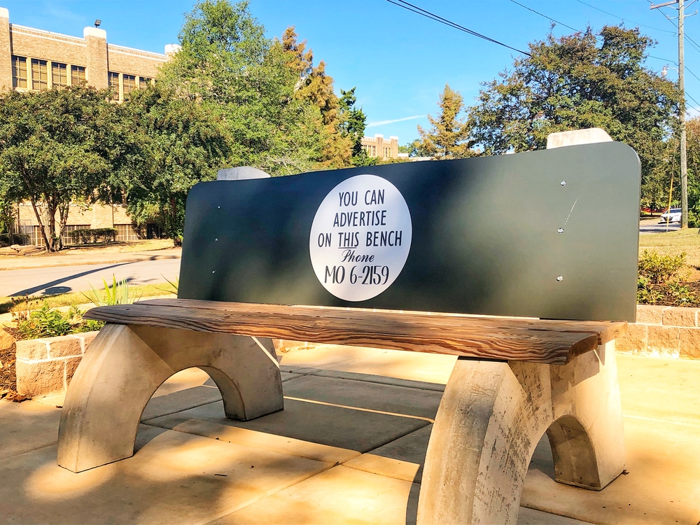 A replica bus bench with concrete curved legs, a wooden seat and black back rest with a circular advertisement reading "You can advertise on this bench - phone MO 6-2159" sits at the corner of 16th Street and S. Park Street. 

At this location, Elizabeth Eckford, one of the Little Rock Nine, waited for a city bus after being turned away by the Arkansas National Guard from entering Central High School and pursued here by an angry mob on Wednesday, September 4, 1957. 

In the background, trees and a sidewalk stand in front of the south wing of Central High School. 
