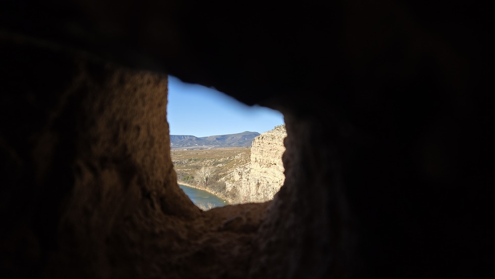 the view out though a small hole in an adobe cliff dwelling wall. A creek is visible below the cliff and mountains are seen in the distance.