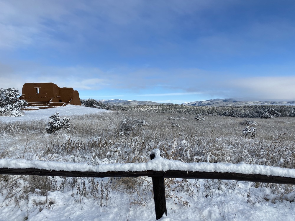 A thick blanket of white snow covers a dark brown fence that closes off a landscape view of the red adobe Spanish Mission and the snowcapped Sangre De Cristo Mountains.