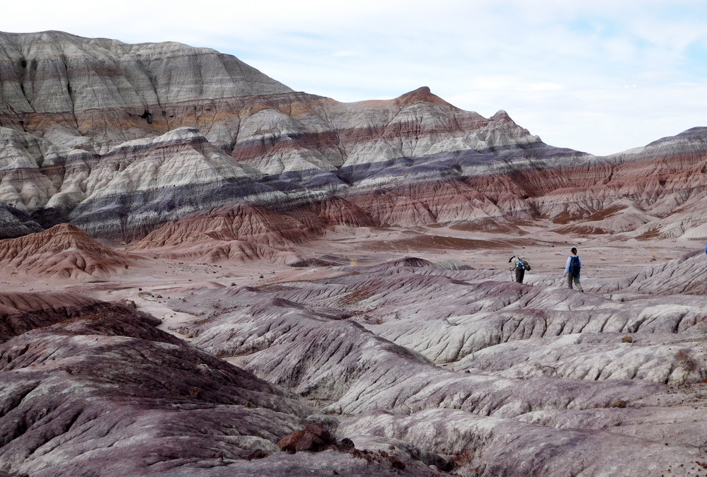 Two hikers in a badland landscape with tilted colorful beds. Partially cloudy sky above.