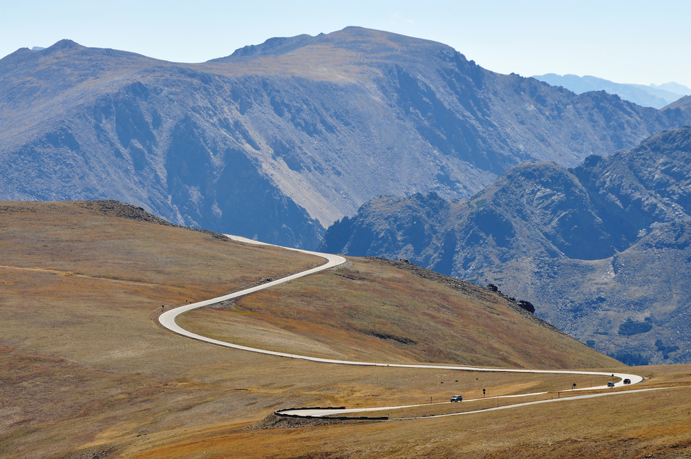 Cars drive a small road dominated by treeless rocky mountains.