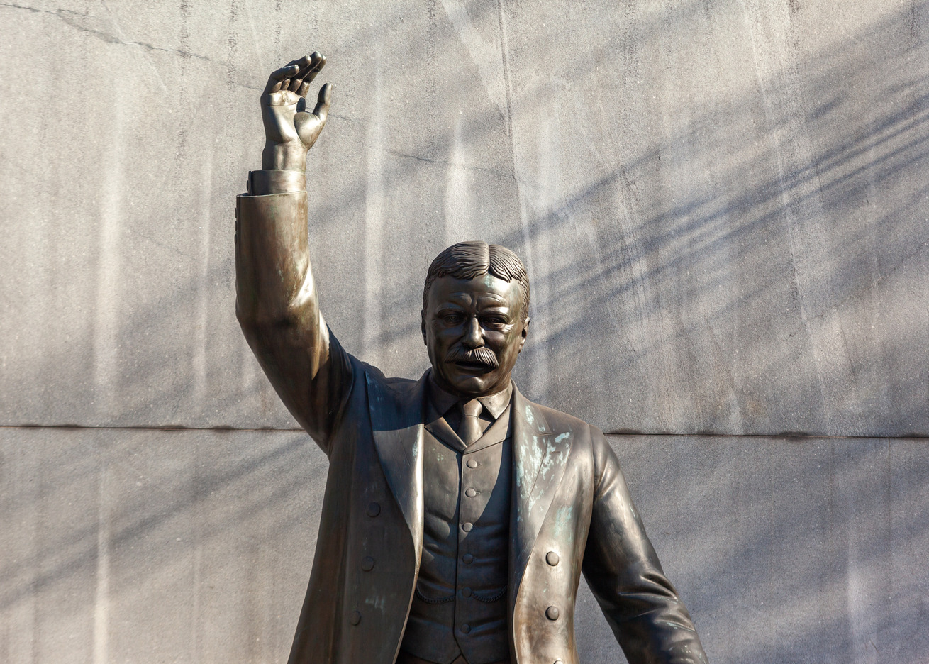 Shadows are cast behind the Theodore Roosevelt Memorial; in which Theodore Roosevelt has his right hand raised and appears to be speaking.