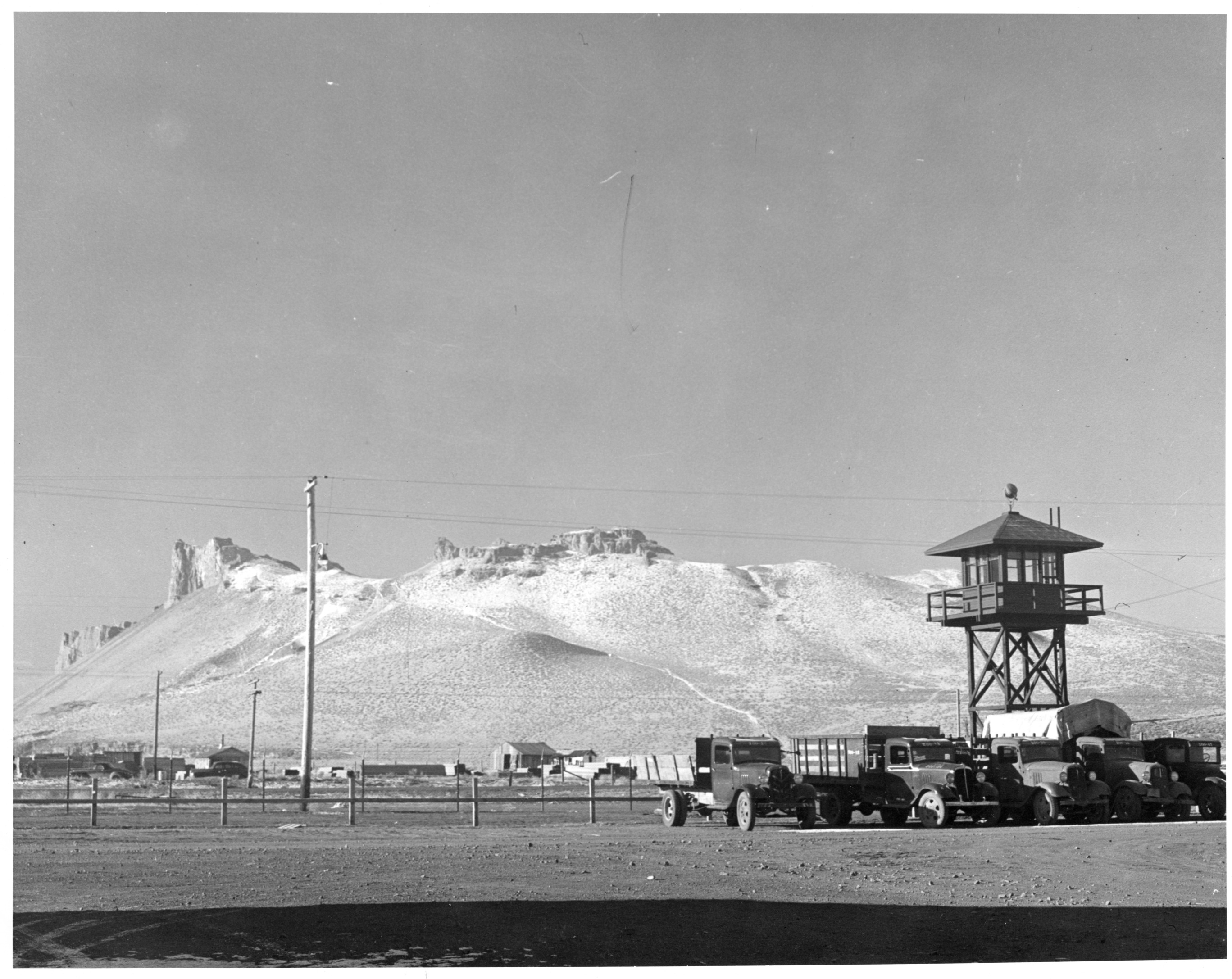 A mountain with distinct ridge formations with flat dirt land in foreground. Four 1930s - 40s era trucks are parked under a wood guard tower.