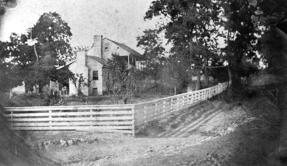 Black and white photo of a two-story frame house surrounded by a white rail fence. 