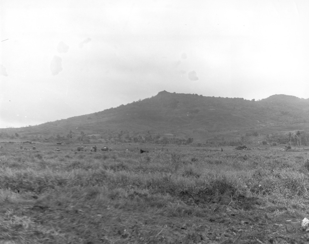A black-and-white landscape photograph shows an expansive grassy plain leading to a large, tree-covered hill under a cloudy sky. Large guns have been set up on the plain.
