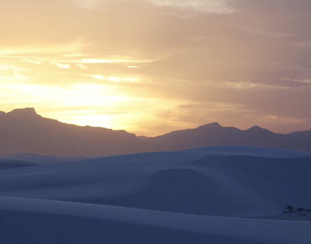 Tall rolling hills of sand dunes at the base of rugged mountains spanning the horizon of the image under a barely lit sunset sky. 